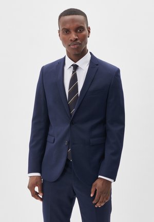 Un homme se tient devant un fond clair, vêtu d’un costume bleu marine, d’une chemise blanche et d’une cravate rayée.