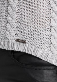 Jasnoszary sweter z dzianiny, z wzorem w warkocze, ribbowym dołem i metalową metką z logo. Połączony z czarnymi spodnimi slim fit.
