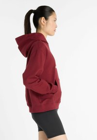 Maroon hoodie with a kangaroo pocket, featuring a hood and ribbed cuffs. Worn with black shorts, shown from the side against a white background.