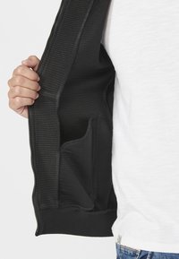 Giacca nera con zip e interno a coste, con tasca visibile. Texture liscia, design aderente e vita elastica. T-shirt bianca sotto.