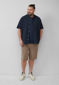 Marineblaues kurzärmliges Button-Down-Hemd, dunkles T-Shirt darunter, hellbraune Shorts und cremefarbene Sneakers. Robustes Material, lockere Passform, funktionales Design.