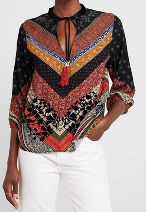 Kobieta w wielobarwnej, wzorzystej bluzce z wiązaniem przy szyi i frędzlami, w białych spodniach, z ręką w kieszeni.