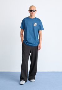 Blå bomulls T-skjorte med "OBEY"-logo, parret med løse svarte bukser og hvite joggesko. Modellen har på seg svarte solbriller og viser arm tatoveringer.