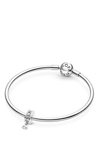 La pulsera de charms de plata de Pandora presenta una banda lisa y redonda, un cierre esférico con letras grabadas y un pequeño charm en forma de corazón con una inscripción.