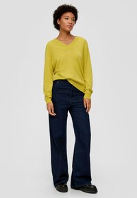 Gelber V-Ausschnitt-Pullover mit langen Ärmeln, kombiniert mit dunkelblauen, weit geschnittenen Jeans und schwarzen, klobigen Schuhen. Glatte Textur, lockere Passform.