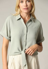 Lichtgroen kortwaardig knoopshirt met een kraag, geknoopt op de taille. Textuurstof met opgerolde mouwen en een ontspannen pasvorm.