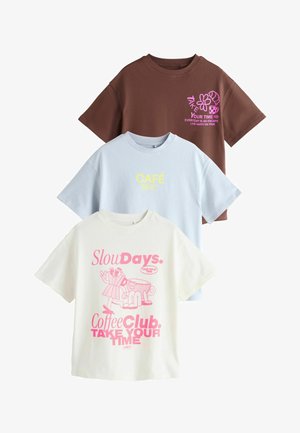 Drei übergroße T-Shirts: braun mit pinkem Grafikdruck, hellblau mit gelbem Text und cremefarben mit pinken Illustrationen und Text; alle aus Baumwolle.