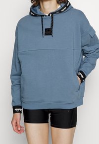 Blå sweatshirt med huva, med svarta dragsnören och logotypficka. Muggarna har svart accent med vit branding. Matcher med svarta shorts.