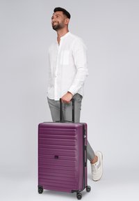 Uomo in camicia bianca e pantaloni grigi sta in piedi tenendo il manico di una grande valigia viola con ruote, guardando verso l'alto e sorridendo.