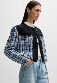 Tweed jakke med et blåt, sort og hvidt ternet mønster. Kort længde med gyldne knaplukninger og frynsede kanter; har to forlommer.
