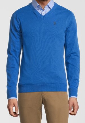 Suéter azul de cuello en V hecho de una tela suave, con puños y dobladillo acanalados. Presenta un logo sutil en el pecho. Llevado sobre una camisa de color azul claro con cuello.