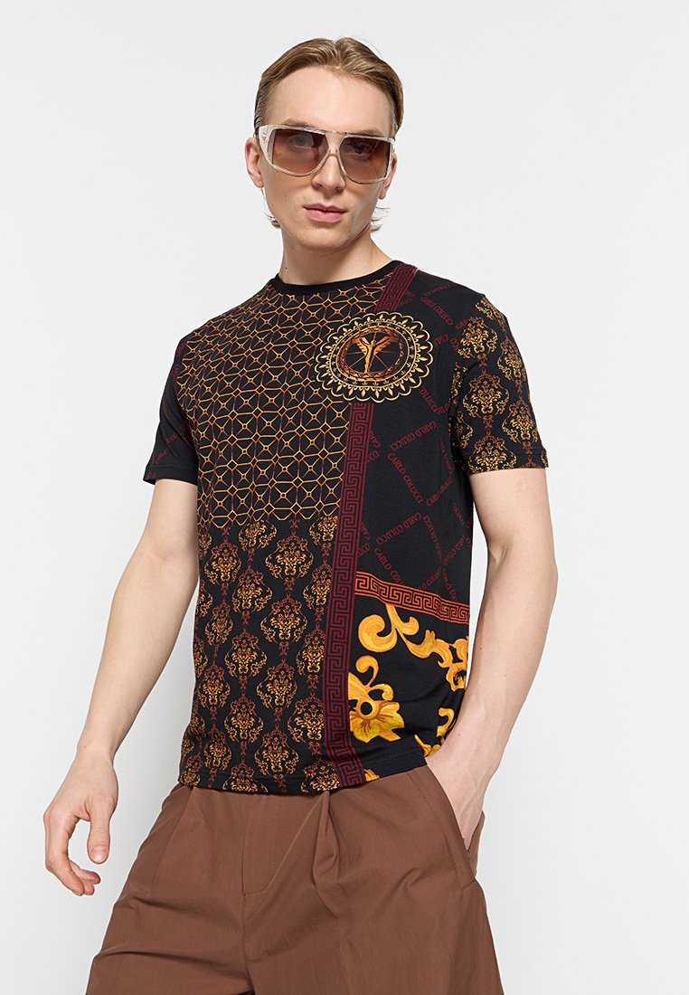Mann trägt übergroße getönte Sonnenbrille, ein schwarzes kurzärmeliges Hemd mit goldenen und roten Mustern und braune Hosen mit einer Hand in der Tasche.