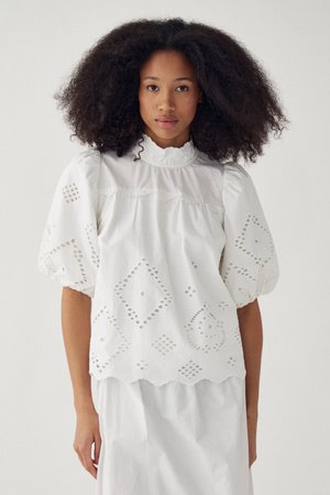 Jeune femme aux cheveux bouclés et volumineux portant un haut blanc à manches bouffantes ajourées et une jupe assortie sur un fond uni.