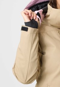 Ærme på beige jakke med sort Velcro-stropdetalje, en hånd justerer det pink og sorte indvendige foer nær kraven.