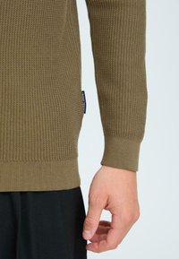 Pulover verde măsliniu cu un model de tricotaj texturat, manșete și tiv cu un model ondulat. Eticheta neagră vizibilă pe lateral. Mână odihnindu-se pe o suprafață din țesătură neagră.
