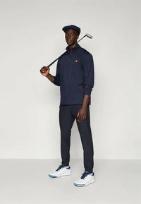 Marineblå polo med lange ermer og glidelås, mørke bukser og hvite joggesko. Holder en golfkølle, iført en matchende marineblå caps med logo.