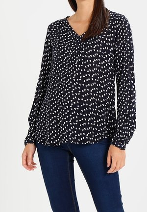 Kobieta ubrana w czarną bluzkę z długim rękawem w białe wzory serc, oraz dopasowane niebieskie dżinsy, stojąca na tle jednolitej białej ściany.