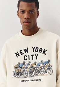 Jeune homme portant un sweat-shirt crème avec un graphisme de six cyclistes en tenue bleue et jaune, ainsi que le texte "NEW YORK CITY" et "1991 / UPDATED GARMENTS".