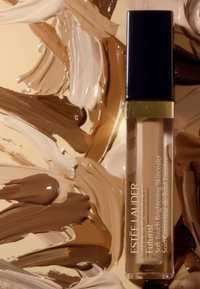 Estee Lauder Futurist Soft Touch Brightening Skincare i en klar tub, med ett marinblått lock och guldaccent, mot en virvlande brun bakgrund.