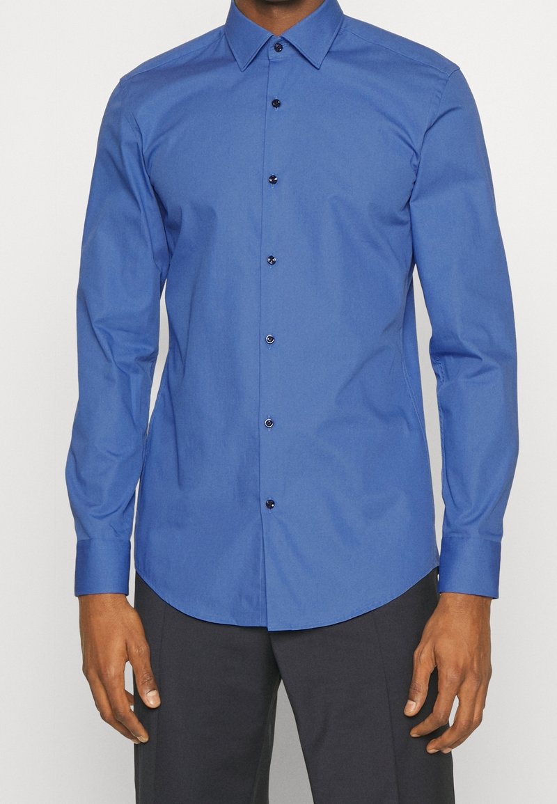 Homme portant une chemise bleue ajustée à manches longues avec des boutons noirs et un pantalon foncé, debout devant un fond blanc.