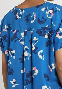 Blue floral blouse featuring a loose fit, short sleeves, and a pleat detail at the back. The fabric has a soft texture with a vibrant floral pattern.