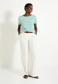 Camiseta rayada verde y blanca de manga corta con botones en los hombros, combinada con pantalones anchos blancos y sandalias marrones.