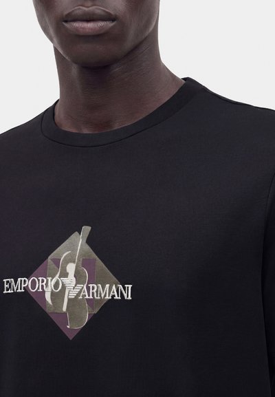 Κοντινό πλάνο ενός ατόμου που φοράει μαύρο μπλουζάκι με γραφικά κιθάρας και το λογότυπο Emporio Armani στο στήθος.