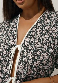 Svart blommönstrad plagg med djup V-ringning, vit knytning och korta ärmar. Har små rosa och gröna blommor överallt.