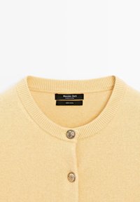 Ljus beige 100% ull kavaj med rund halsringning, med ribbad fåll och två silverfärgade knappstängningar framtill.