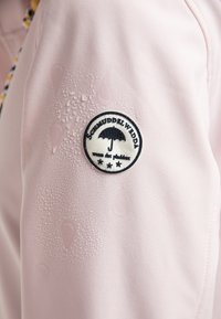 Veste waterproof rose clair avec un écusson circulaire noir et blanc, montrant des gouttes d'eau sur la surface du tissu.