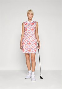 Ermeløs golfkjole i rosa, lilla og hvitt abstrakt mønster, med krage og innsvingt midje. Kombinert med hvite sko og golfkølle.