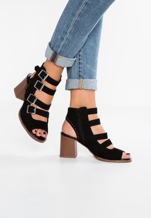 Sorte ruskind open-toe sandaler med fire justerbare stropper, lynlås i siden og en blokhæl med en struktureret, brun finish.