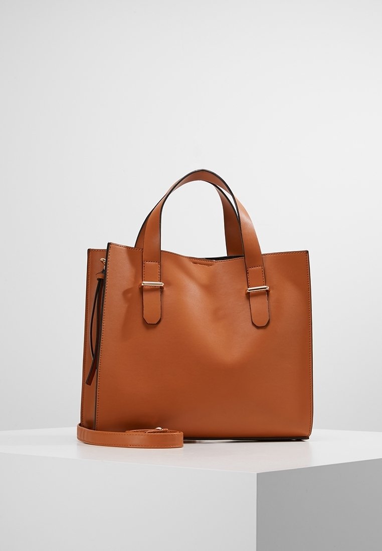 Sac fourre-tout en cuir marron avec deux poignées supérieures et une bandoulière amovible. Texture lisse, design minimaliste, sans motifs visibles.