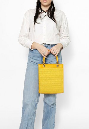 Vrouw draagt een witte blouse en lichtblauwe spijkerbroek, houdt een gele getextureerde tas met ronde handvatten vast, voor een witte achtergrond.