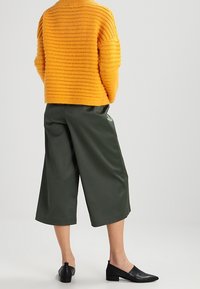 Maglione lavorato a maglia spessa di un brillante arancione con un motivo testurizzato, abbinato a pantaloni cropped larghi di un verde scuro e scarpe basse nere.