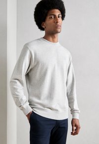 Light gray sweatshirt with long sleeves, ribbed cuffs and hem. Features a small blue logo on the chest. Paired with dark blue pants.