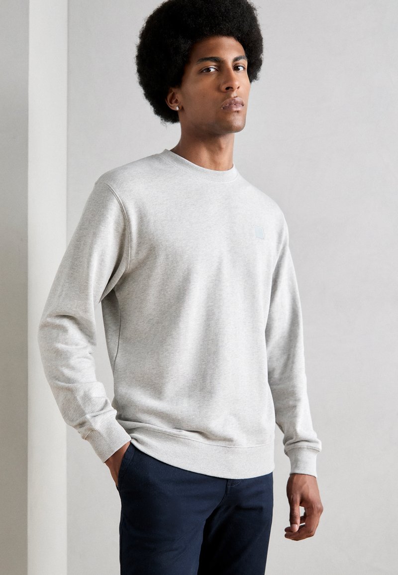 Light gray sweatshirt with long sleeves, ribbed cuffs and hem. Features a small blue logo on the chest. Paired with dark blue pants.