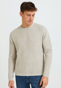 Helle beige Langarm-Sweater aus strukturiertem Material mit rundem Halsausschnitt und kontrastierenden Besätzen, kombiniert mit blauen Jeans.