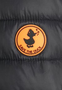 Orange cirkulært mærke med en sort and-silhuet og musiknoter, mærket "SAVE THE DUCK" på sort quiltet stof.