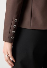 Hnědá saka na míru s rukávem se čtyřmi stříbrnými knoflíky. Tkanina vypadá hladce, vedle černého oděvu. Celkový design je přiléhavý.