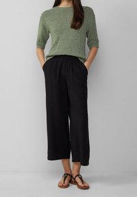 Femme portant un haut en tricot vert à manches courtes, un pantalon large noir cropped et décontracté, ainsi que des sandales à lanières noires, se tenant devant un fond uni.