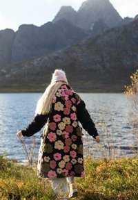 Čierny dlhý kabát s kvetinovým vzorom, zdobený viacfarebnými kvetmi. Kabát má jemnú textúru a je skombinovaný s ružovou čiapkou a svetlými nohavicami.