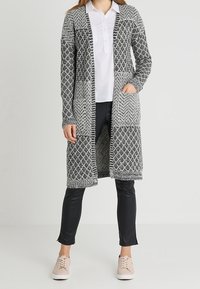 Cárdigan largo de punto en blanco y negro con patrones geométricos, con apertura en la parte delantera, bolsillos y puños de canalé; combinado con pantalones negros y zapatillas rosas.