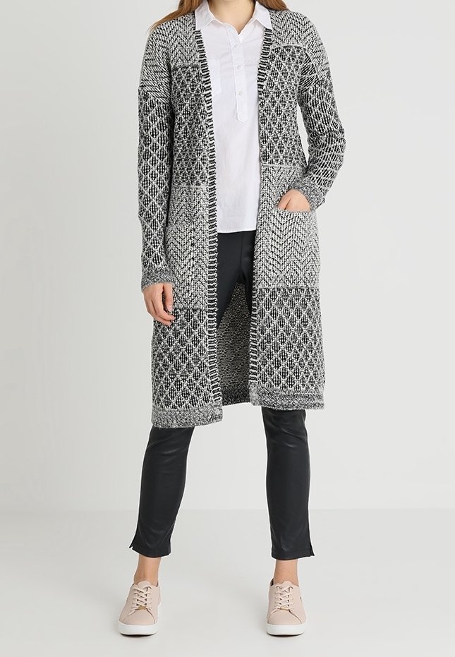 Cárdigan largo de punto en blanco y negro con patrones geométricos, con apertura en la parte delantera, bolsillos y puños de canalé; combinado con pantalones negros y zapatillas rosas.