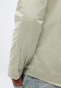 Άνδρας που φοράει πουκάμισο με κάθετες ρίγες ανοιχτού πράσινου και λ ευκού, με μακριά μανίκια, μπαλωμένο σε ανοιχτόχρωμα μπλε τζιν, με το χέρι του να είναι μερικώς στην τσέπη.