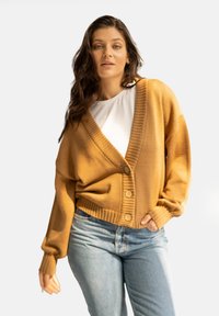 Musztardowy kardigan z dekoltem w serek, w prążkowanej strukturze, z pięcioma guzikami z przodu. Nosi się go na białej koszuli, w zestawieniu z niebieskimi dżinsami.