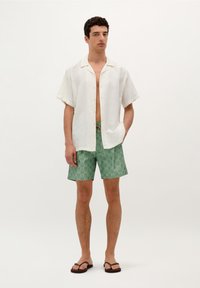 Chemise blanche à manches courtes avec un motif textur�é, associée à un short à motifs verts et des tongs marron, se tenant contre un fond clair.