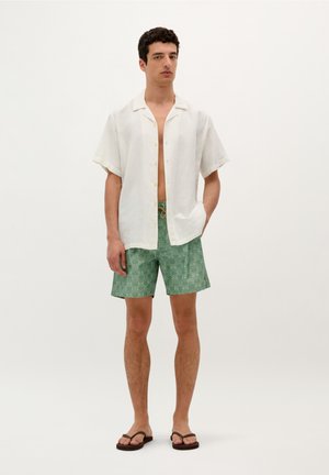 Weißes Kurzarmhemd mit strukturiertem Muster, kombiniert mit grünem gemustertem Shorts und braunen Flip-Flops, steht vor einem hellen Hintergrund.