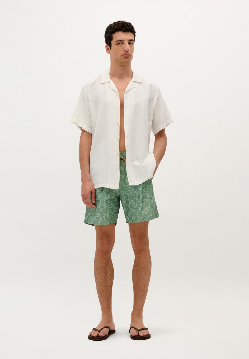 Chemise blanche à manches courtes avec un motif texturé, associée à un short à motifs verts et des tongs marron, se tenant contre un fond clair.