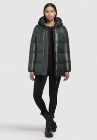 Donkergroene pufferjack met capuchon, met een boxy ontwerp, glanzend materiaal, knoop- en ritsafsluitingen, en uitgesproken gestikte secties.